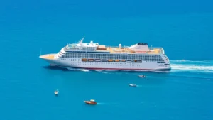 Large modern cruise ship sailing through turquoise Mediterranean waters with smaller fishing boats nearby, showing scale difference and marine traffic density in popular tourist destinations